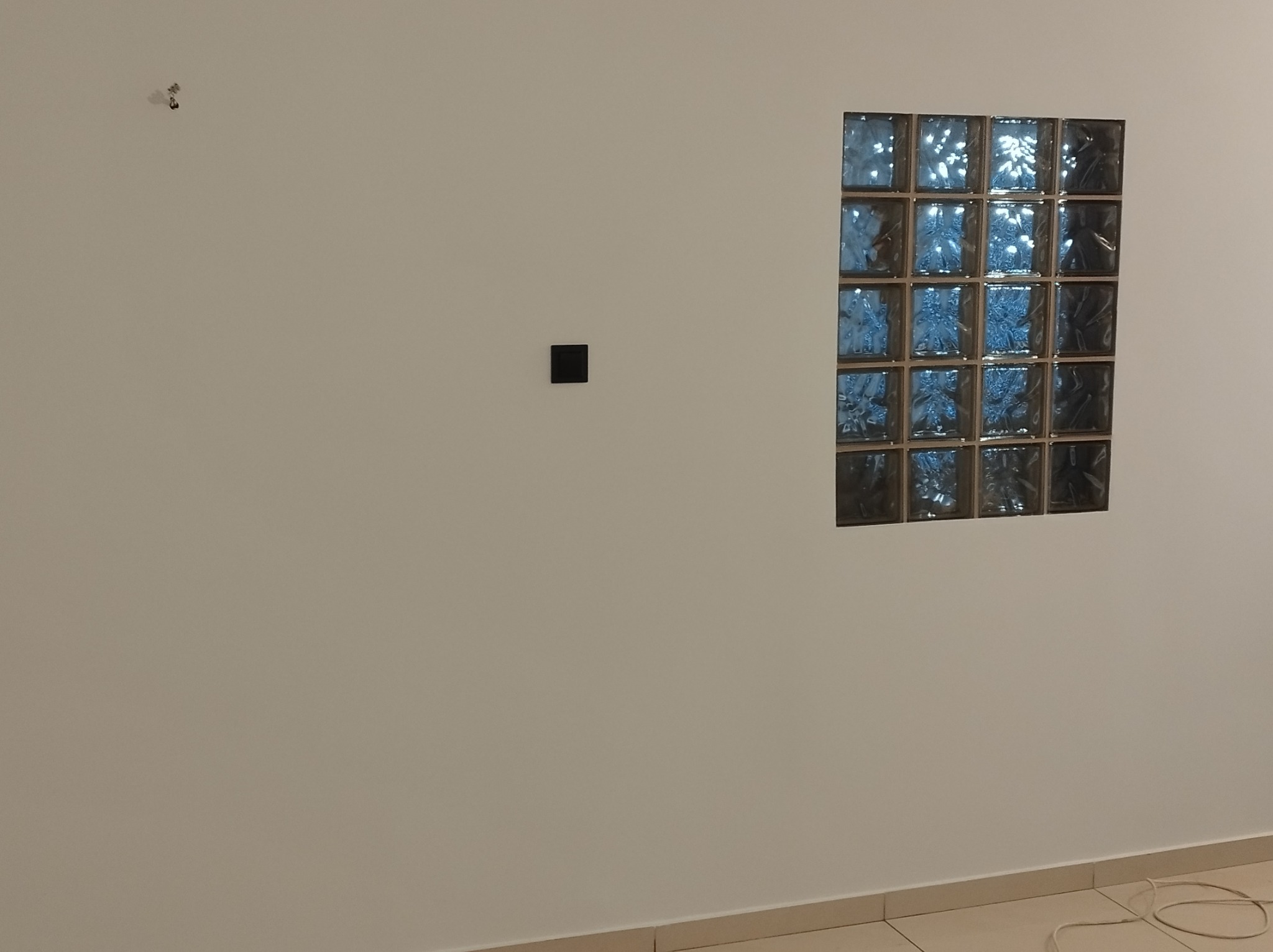 Ściana z zamontowanym panelem z luksferów w kształcie kwadratu, z widocznym gniazdkiem elektrycznym i śladami po wierceniu, ukazująca etap prac wykończeniowych.