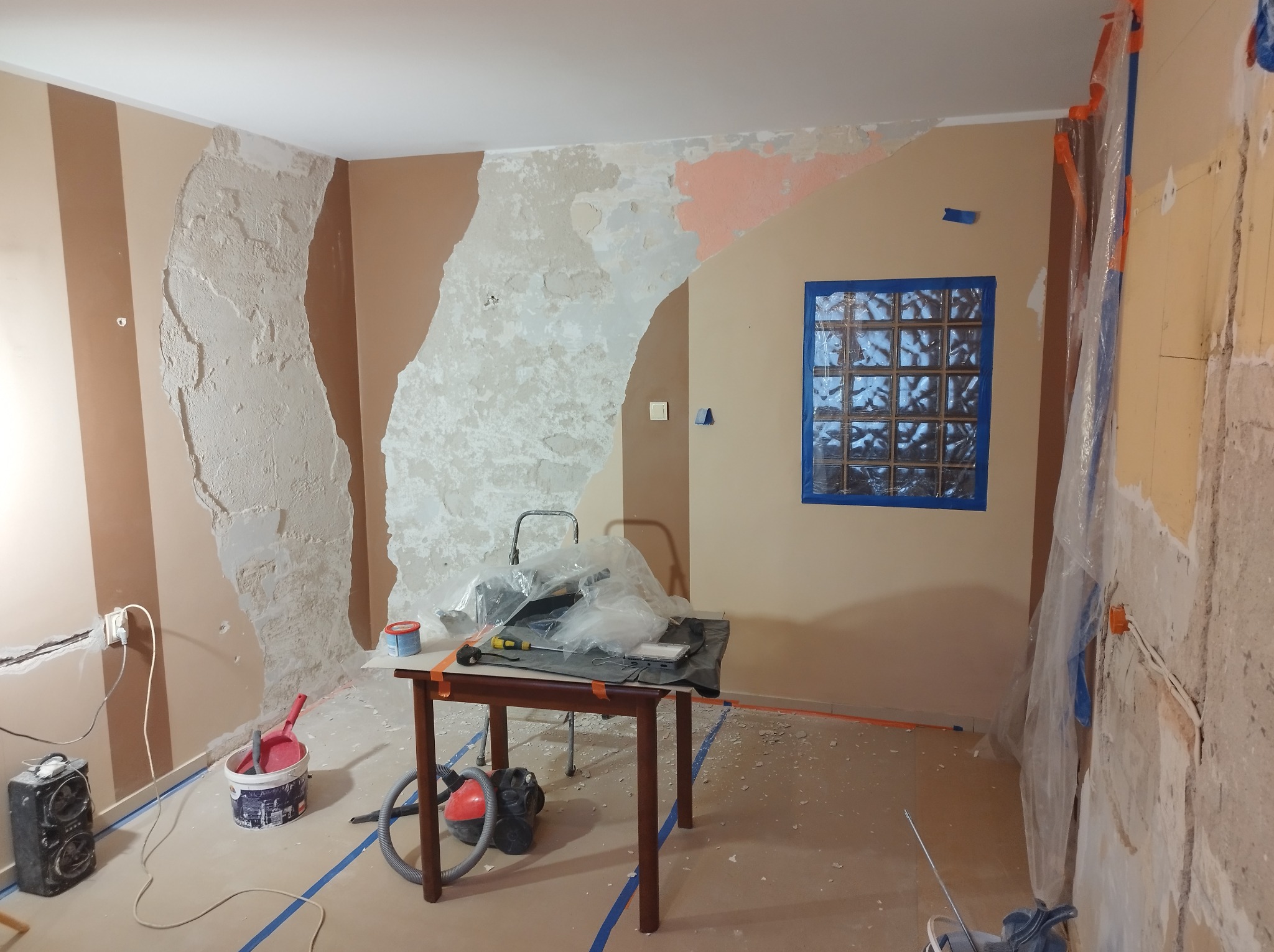 Pokój w trakcie przygotowania do prac wykończeniowych: częściowo usunięta stara farba, zabezpieczone okno z luksferów taśmą malarską, widoczne narzędzia i akcesoria na stole oraz odkurzacz i wiadro...