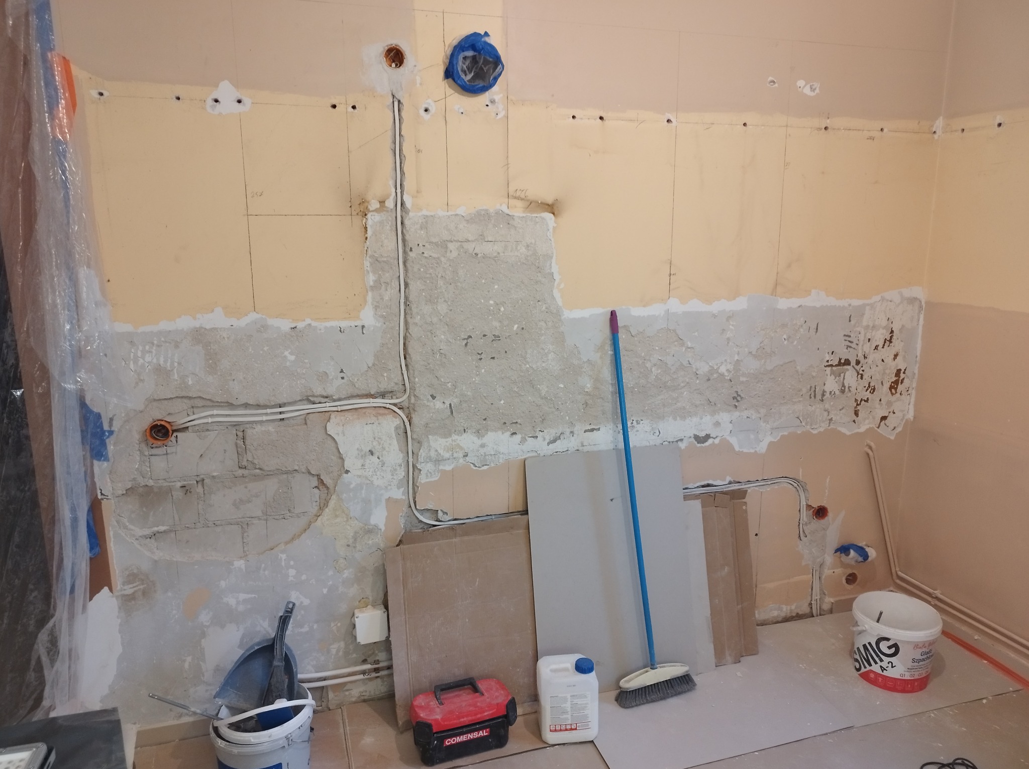 Fragment ściany w trakcie prac przygotowawczych do remontu: odsłonięte przewody elektryczne, częściowo usunięta stara farba, widoczne ślady po szpachlowaniu, narzędzia pracy i materiały budowlane...