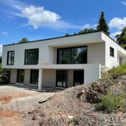 Nowoczesny, dwukondygnacyjny dom z płaskim dachem w trakcie wykończenia elewacji zewnętrznej, widoczny z perspektywy terenu z nasypem ziemi i gruzu, z czarnymi ramami okiennymi.