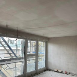 Kamil Laskowski-usługi budowlane - Pokój w trakcie remontu, ściany i sufit pokryte świeżo nałożoną gładzią, widoczne wystające przewody elektryczne i fragment rusztowania za oknem.