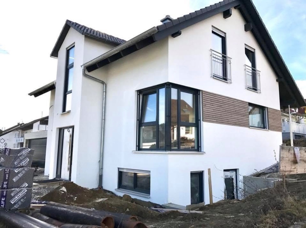 Biały dom jednorodzinny z szarą dachówką i czarnymi ramami okiennymi, częściowo wykończony z elementami drewnianej elewacji, w trakcie prac wykończeniowych, widoczne materiały budowlane i rury