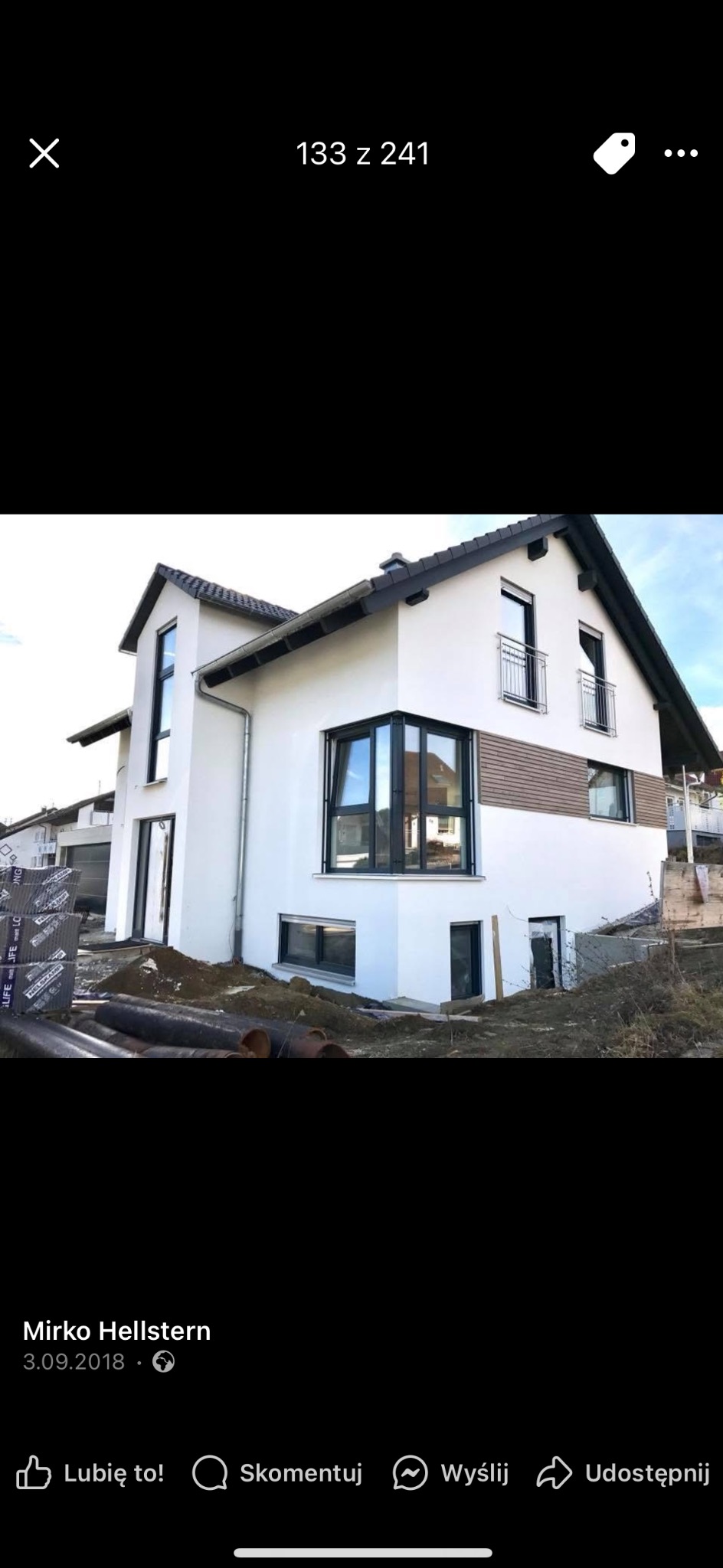 Biały dom w trakcie wykończenia, widoczne rury i materiały budowlane na posesji, elewacja częściowo wykończona drewnem.