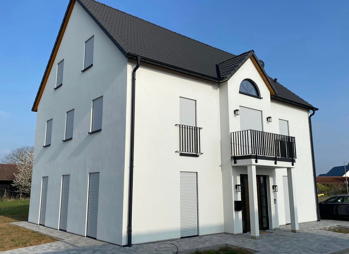 Nowoczesny, dwupiętrowy dom z białymi ścianami zewnętrznymi, czarnym dachem, balkonami z czarnymi balustradami i szarymi roletami okiennymi, sfotografowany w słoneczny dzień.