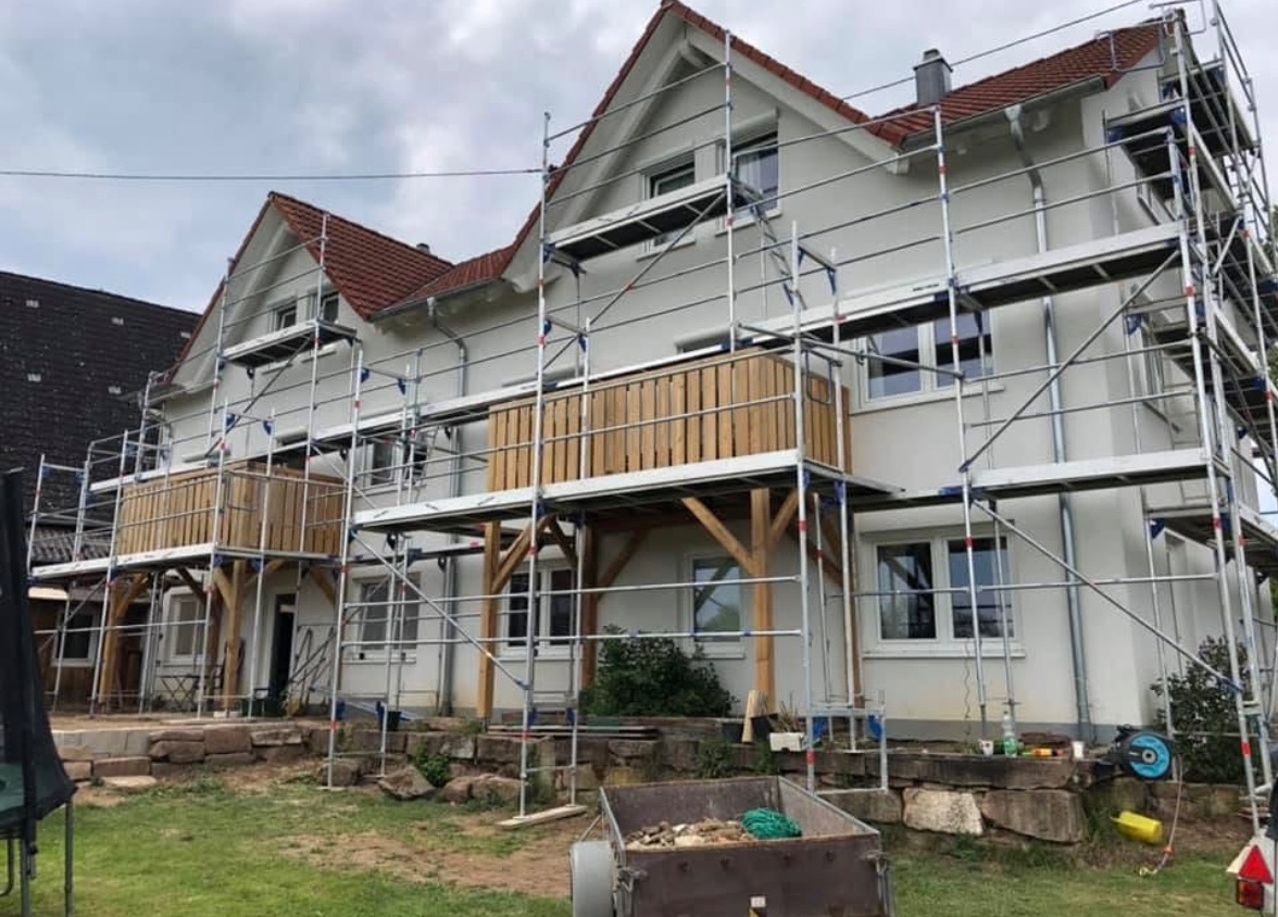 Dom jednorodzinny w trakcie remontu elewacji z użyciem rusztowania, widoczne drewniane balustrady balkonów i przyczepka z gruzem