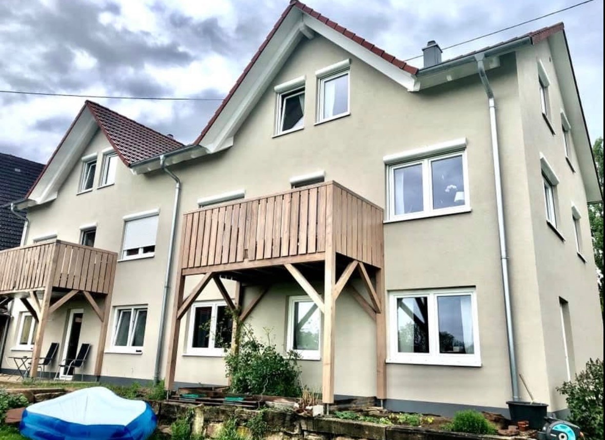 Odnowiona elewacja dwupiętrowego domu z drewnianymi balkonami, widoczne okna z roletami zewnętrznymi i nowa instalacja rynnowa.