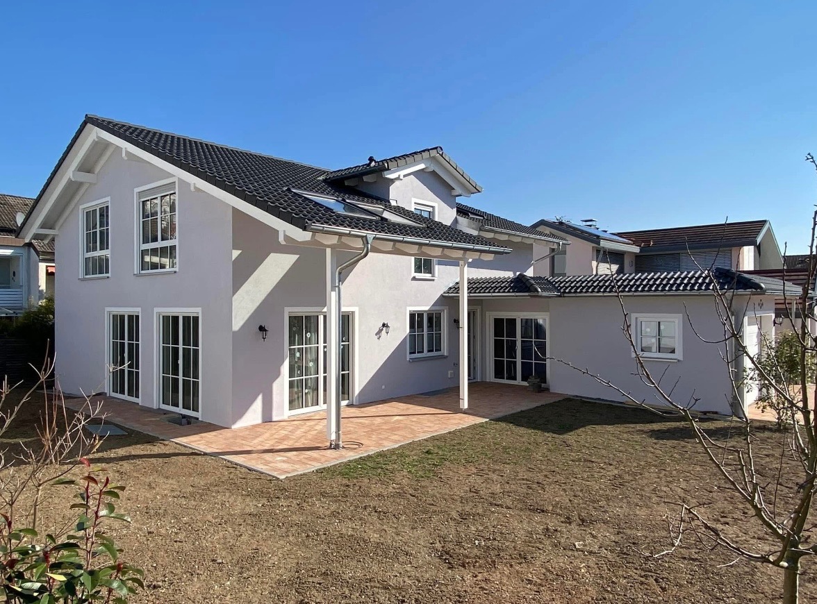 Jasnoszary dom jednorodzinny z ciemną dachówką i tarasem z cegły klinkierowej, widok z ogrodu w słoneczny dzień.