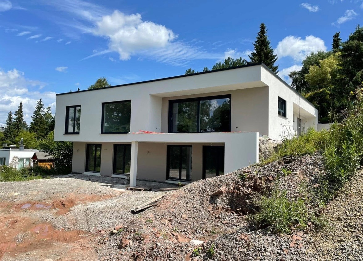 Nowoczesny, dwukondygnacyjny dom z płaskim dachem w trakcie wykończenia elewacji zewnętrznej, widoczny z perspektywy terenu z nasypem ziemi i gruzu, z czarnymi ramami okiennymi.