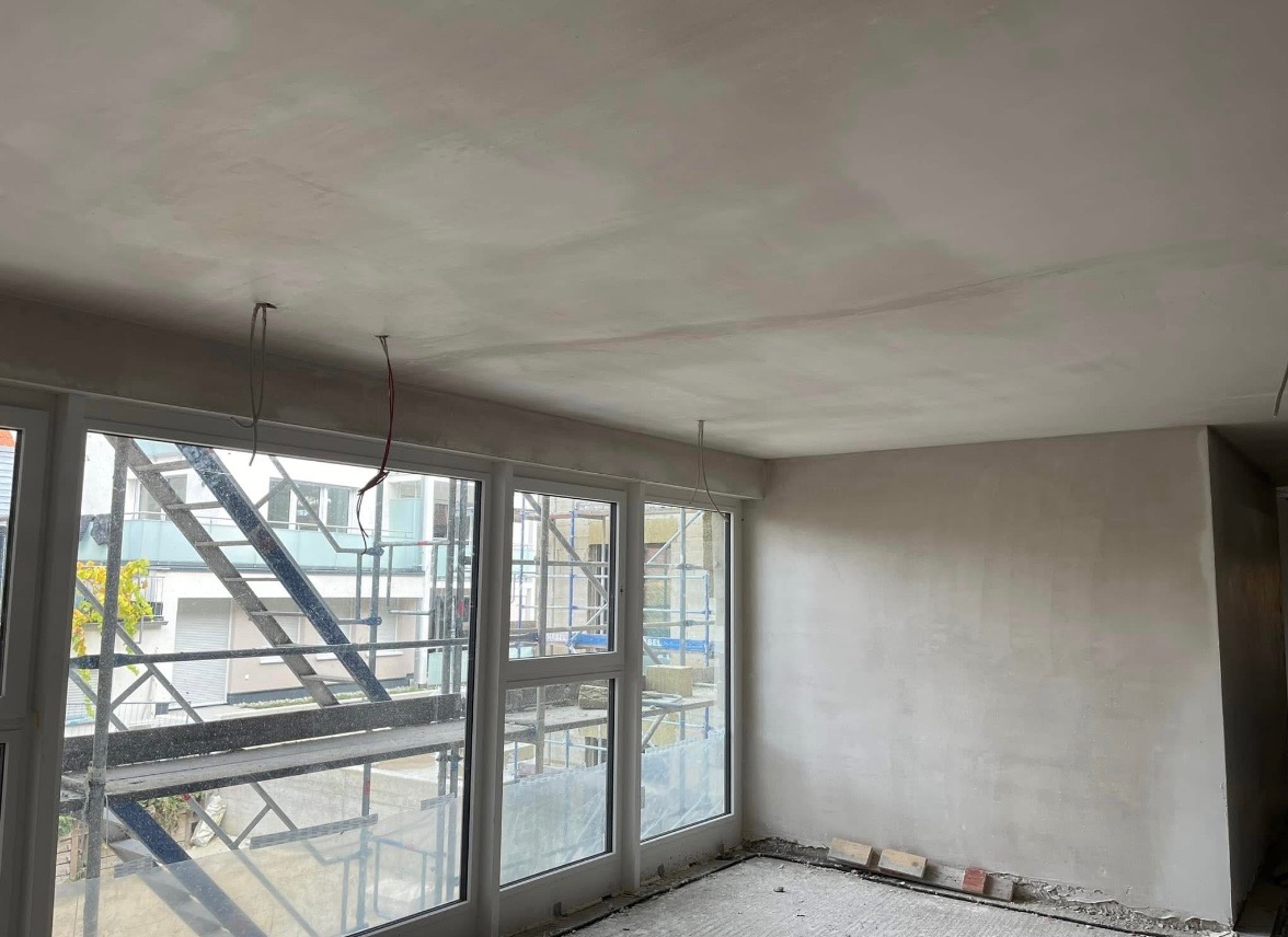 Pokój w trakcie remontu, ściany i sufit pokryte świeżo nałożoną gładzią, widoczne wystające przewody elektryczne i fragment rusztowania za oknem.