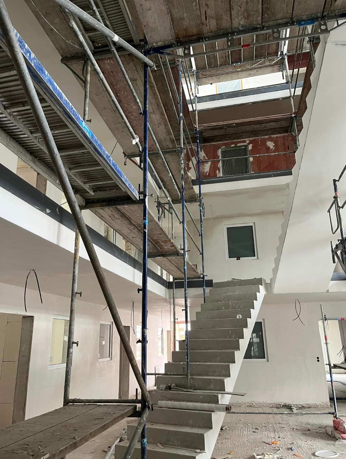 Wnętrze w trakcie prac wykończeniowych: surowe schody betonowe, rusztowania z drewnianymi pomostami, ściany częściowo otynkowane, widoczne instalacje elektryczne, okna wstawione, podłoga...