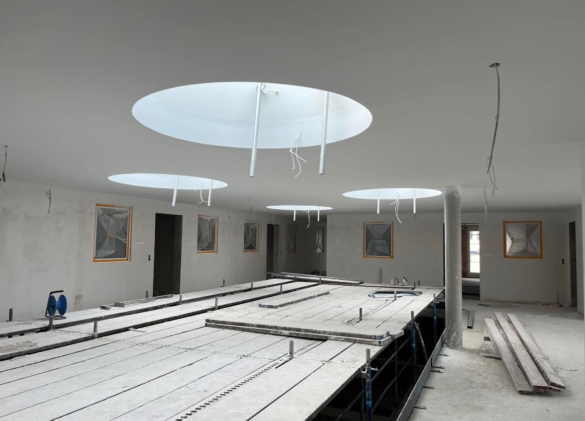 Wnętrze w trakcie prac wykończeniowych z widocznymi rusztowaniami, przygotowanymi ścianami pod malowanie, zabezpieczonymi oknami i okrągłymi świetlikami w suficie.