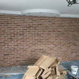 Ściana wyłożona cegłą dekoracyjną w trakcie prac wykończeniowych, widoczne kartony, drabina i zabezpieczenia podłogi.