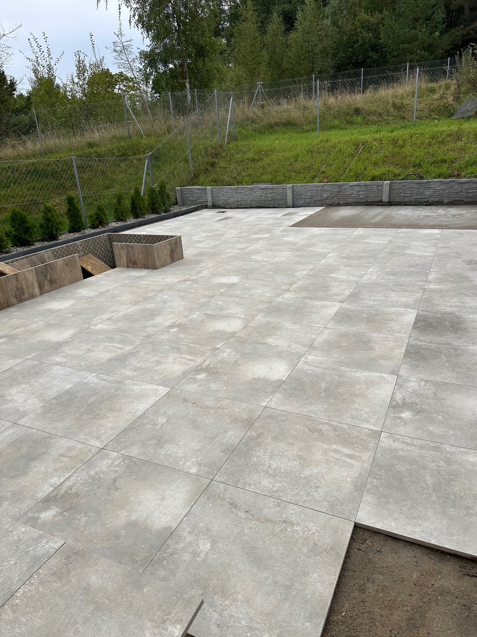 Układanie szarych płyt tarasowych na zewnątrz, widoczne niedokończone fragmenty i przygotowane podłoże, w tle trawnik i ogrodzenie.