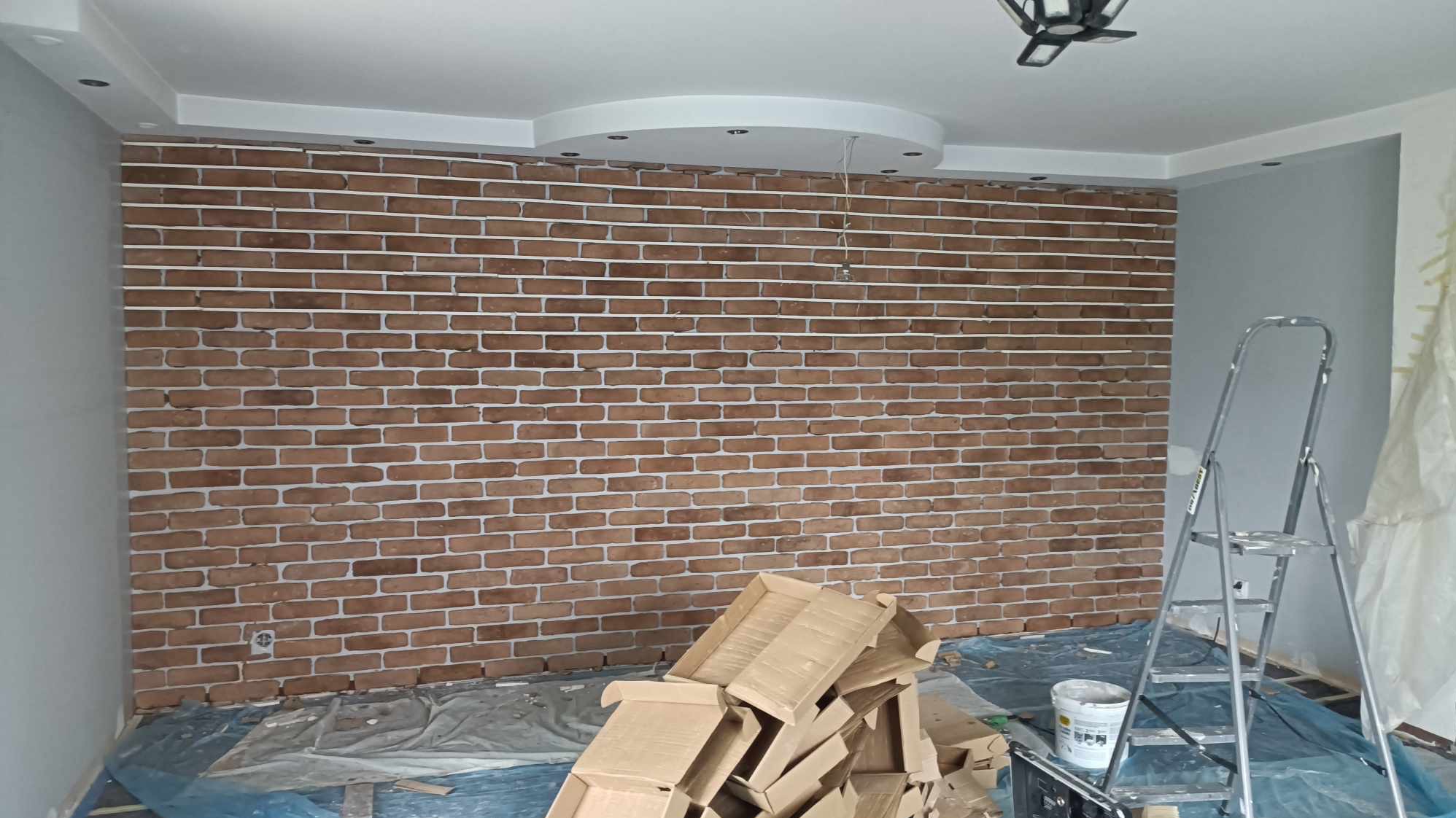 Ściana wyłożona cegłą dekoracyjną w trakcie prac wykończeniowych, widoczne kartony, drabina i zabezpieczenia podłogi.