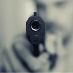 Czarno-białe ujęcie lufy pistoletu skierowanej w stronę widza, z rozmytym tłem postaci.