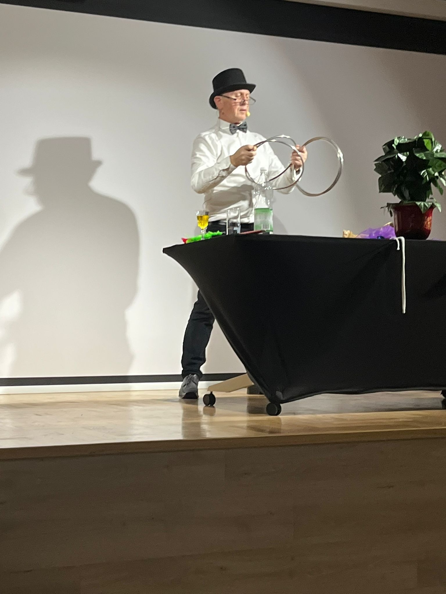 Iluzjonista w cylindrze i muszce prezentuje trik z metalowymi obręczami na scenie. W tle cień kapelusza na ścianie i stół z rekwizytami.