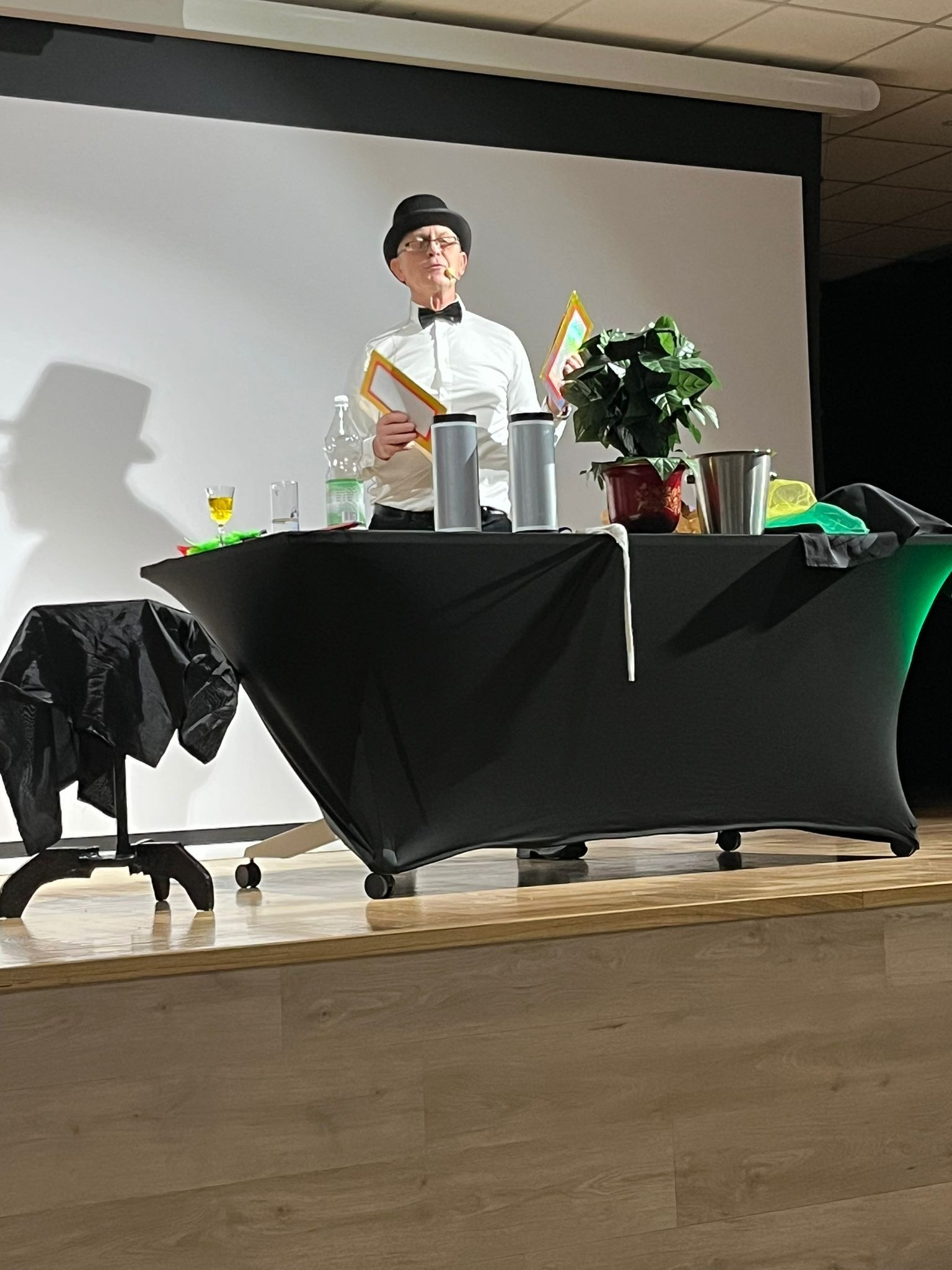 Ustawiony na scenie iluzjonista w cylindrze i muszce, trzymający rekwizyty. Na stole akcesoria, roślina doniczkowa i czarny materiał. W tle biały ekran.