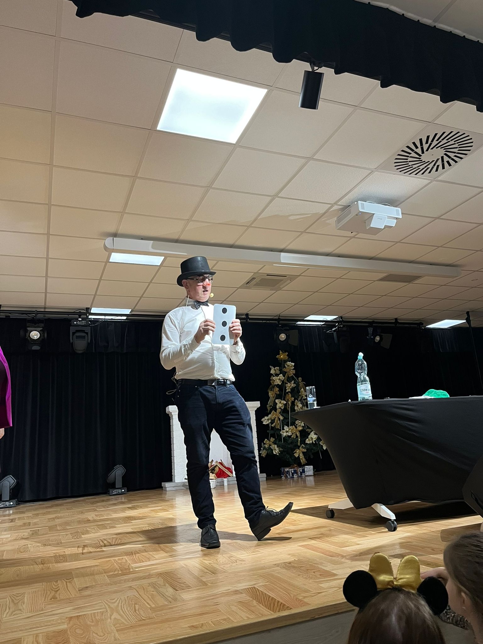 Uśmiechnięty iluzjonista w cylindrze prezentuje kartę z trzema kropkami na scenie z dekoracjami. W tle czarna kurtyna i oświetlenie sceniczne.