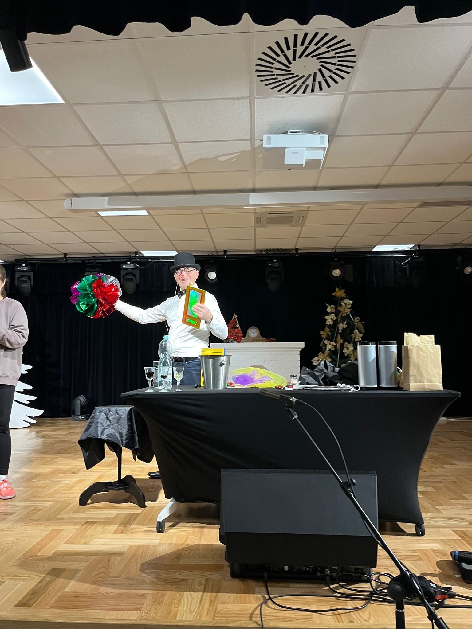 Uśmiechnięty mężczyzna w cylindrze prezentuje rekwizyty: kolorową kulę i ramkę, na scenie z czarnym tłem i stołem z akcesoriami. Oświetlenie sceniczne.