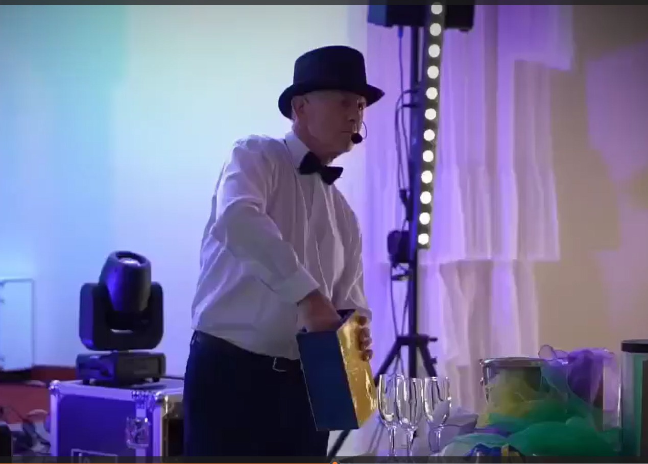 Iluzjonista w cylindrze i muszce, w trakcie pokazu z rekwizytami na scenie, oświetlony reflektorami. W tle białe zasłony i sprzęt oświetleniowy.