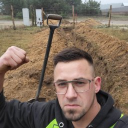 Mateusz Nowakowski - Mężczyzna w okularach, z zaciśniętą pięścią, stoi nad wykopem, obok wbita łopata, w tle widoczne drzewa i słupki ogrodzenia.