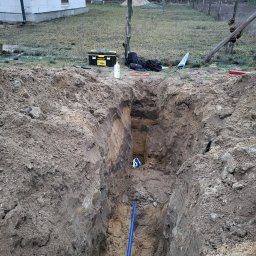 Mateusz Nowakowski - Głęboki wykop w ziemi z niebieską rurą wodociągową na dnie, widoczny fragment instalacji, obok wykopu narzędzia i skrzynka Stanley, w tle budynek w stanie surowym i las.