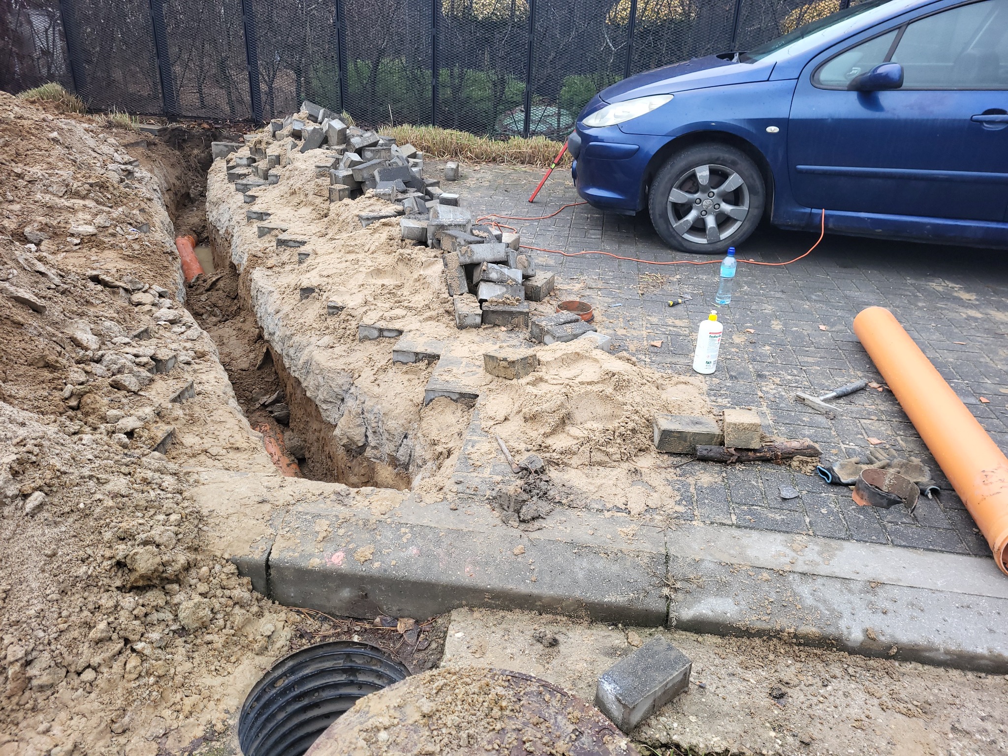 Wykop w ziemi z pomarańczową rurą kanalizacyjną, obok sterta szarych kostek brukowych, niebieski samochód i narzędzia na chodniku.
