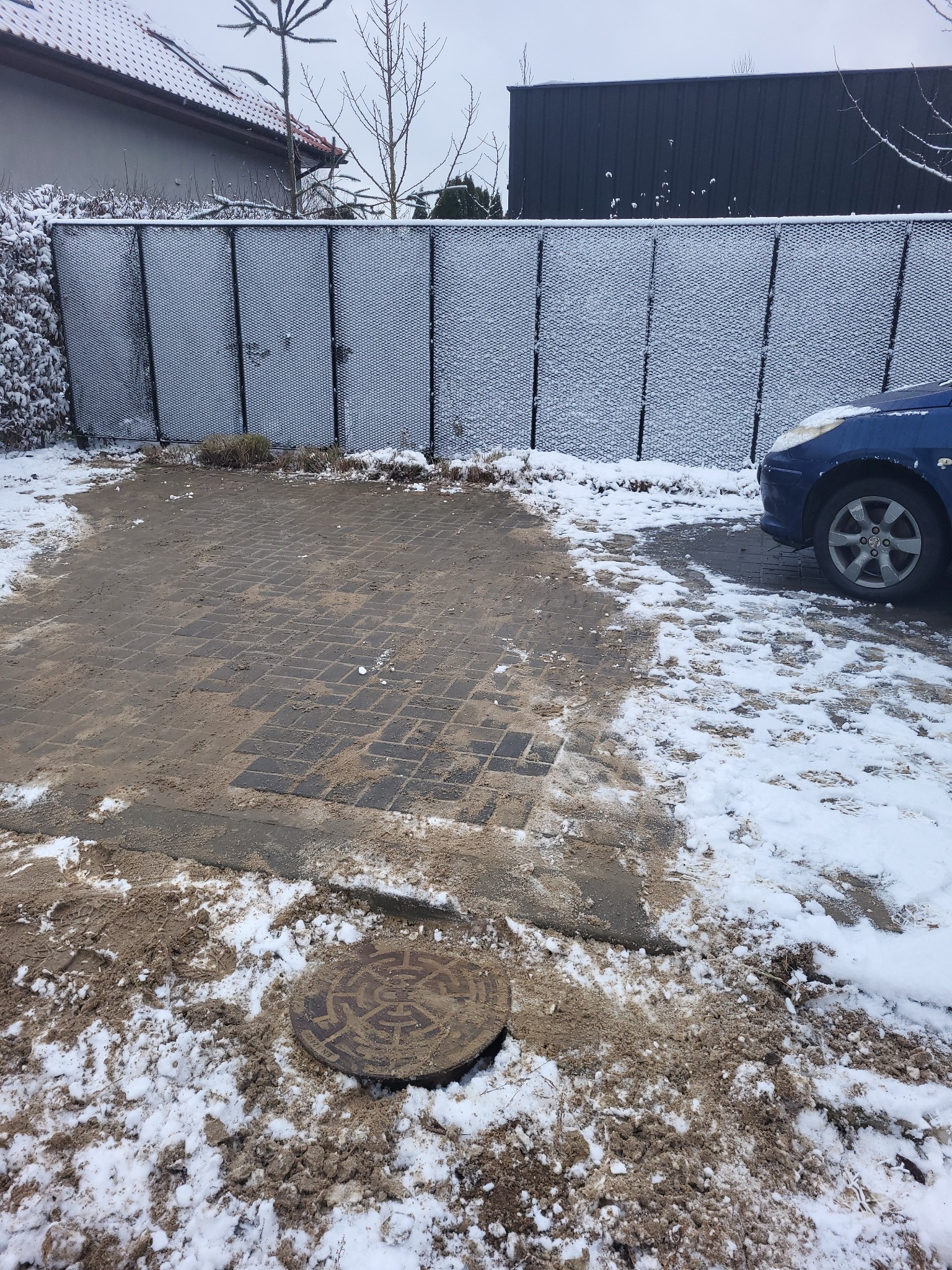 Zewnętrzna studzienka kanalizacyjna na wybrukowanym podjeździe, częściowo pokrytym śniegiem; w tle ogrodzenie z siatki i zaparkowany samochód.