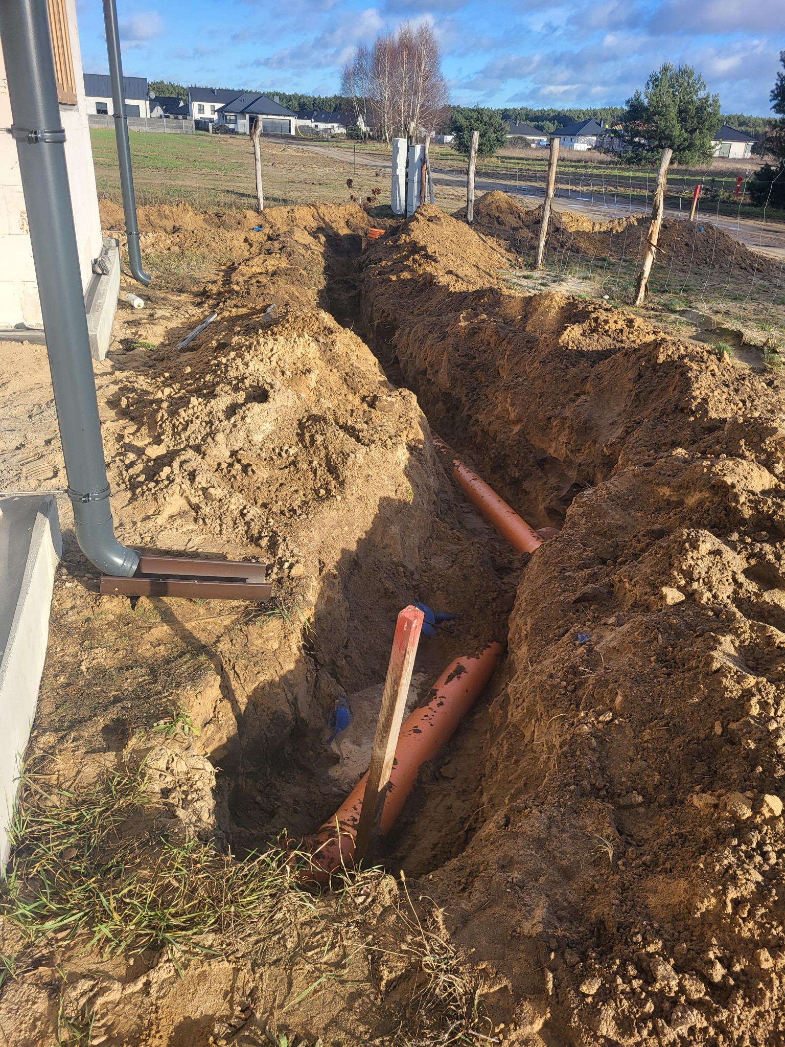 Wykop w ziemi z pomarańczowymi rurami ułożonymi w wykopie, widoczne elementy instalacji przy budynku, w tle ogrodzenie i zabudowa jednorodzinna.