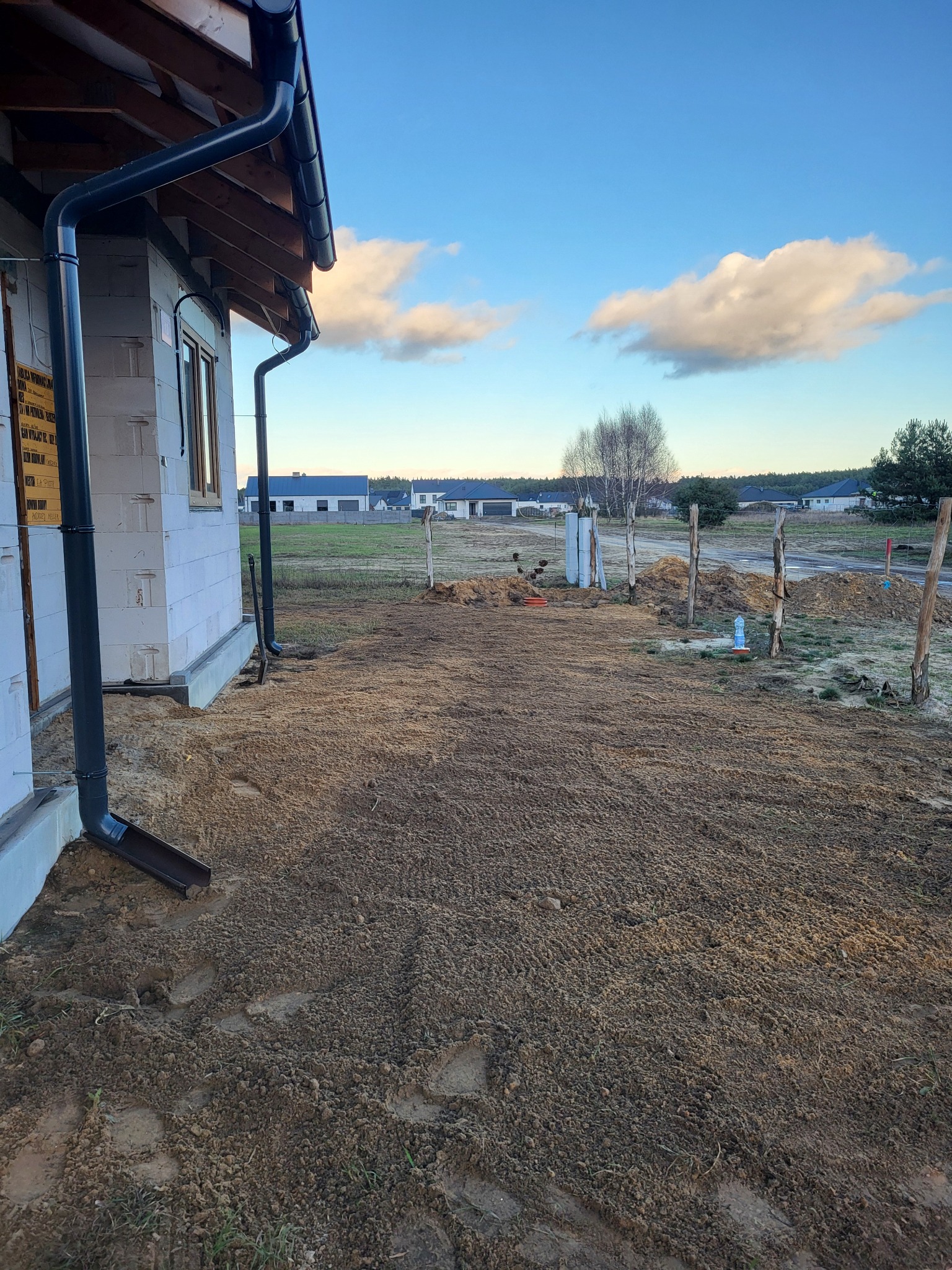 Nowy dom w stanie surowym z zamontowanymi rynnami, przygotowany teren pod trawnik, widoczne słupki ogrodzeniowe i butelka wody.