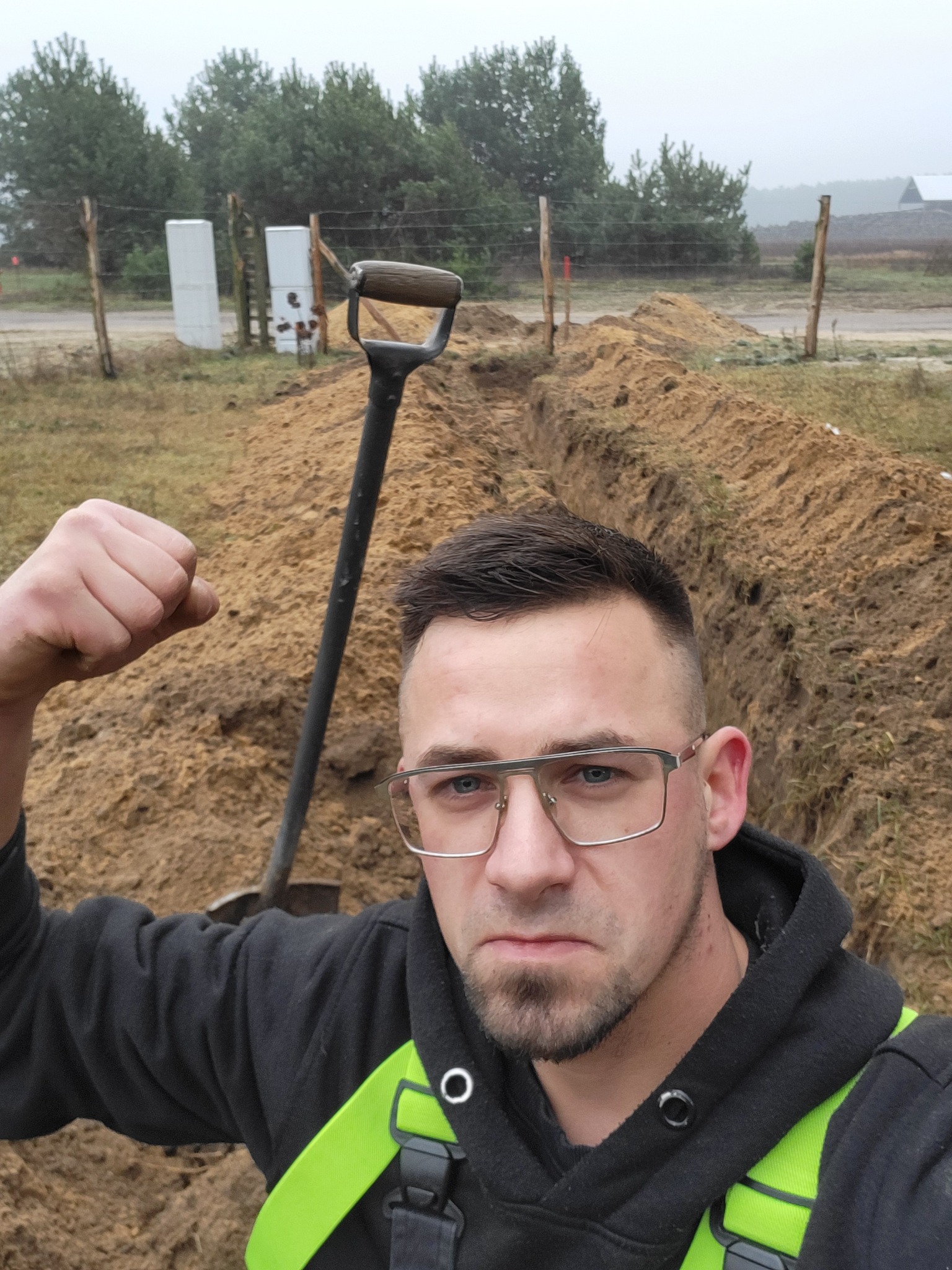 Mężczyzna w okularach, z zaciśniętą pięścią, stoi nad wykopem, obok wbita łopata, w tle widoczne drzewa i słupki ogrodzenia.