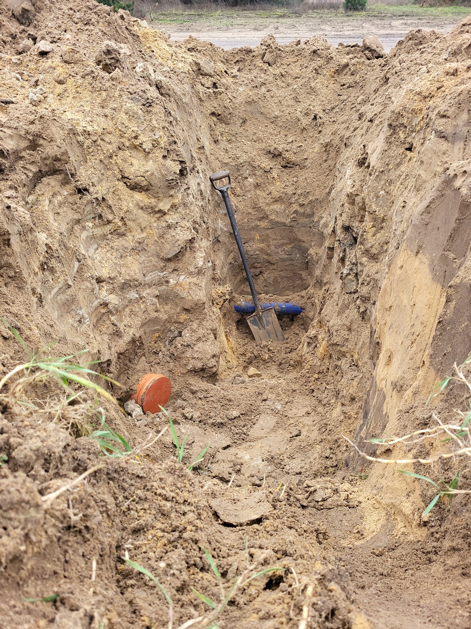 Głęboki wykop w ziemi z widoczną rurą kanalizacyjną, łopatą opartą o ścianę wykopu i pomarańczową zaślepką z boku.