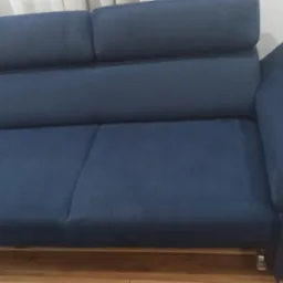 Ciemnoniebieska kanapa z regulowanymi zagłówkami i metalowymi nóżkami na drewnianej podłodze, obok widoczny biały robot sprzątający.