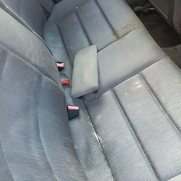 Picuś - Brudna, szara tylna kanapa samochodowa z widocznymi plamami i okruchami, z podłokietnikiem i czerwonymi elementami pasów bezpieczeństwa.