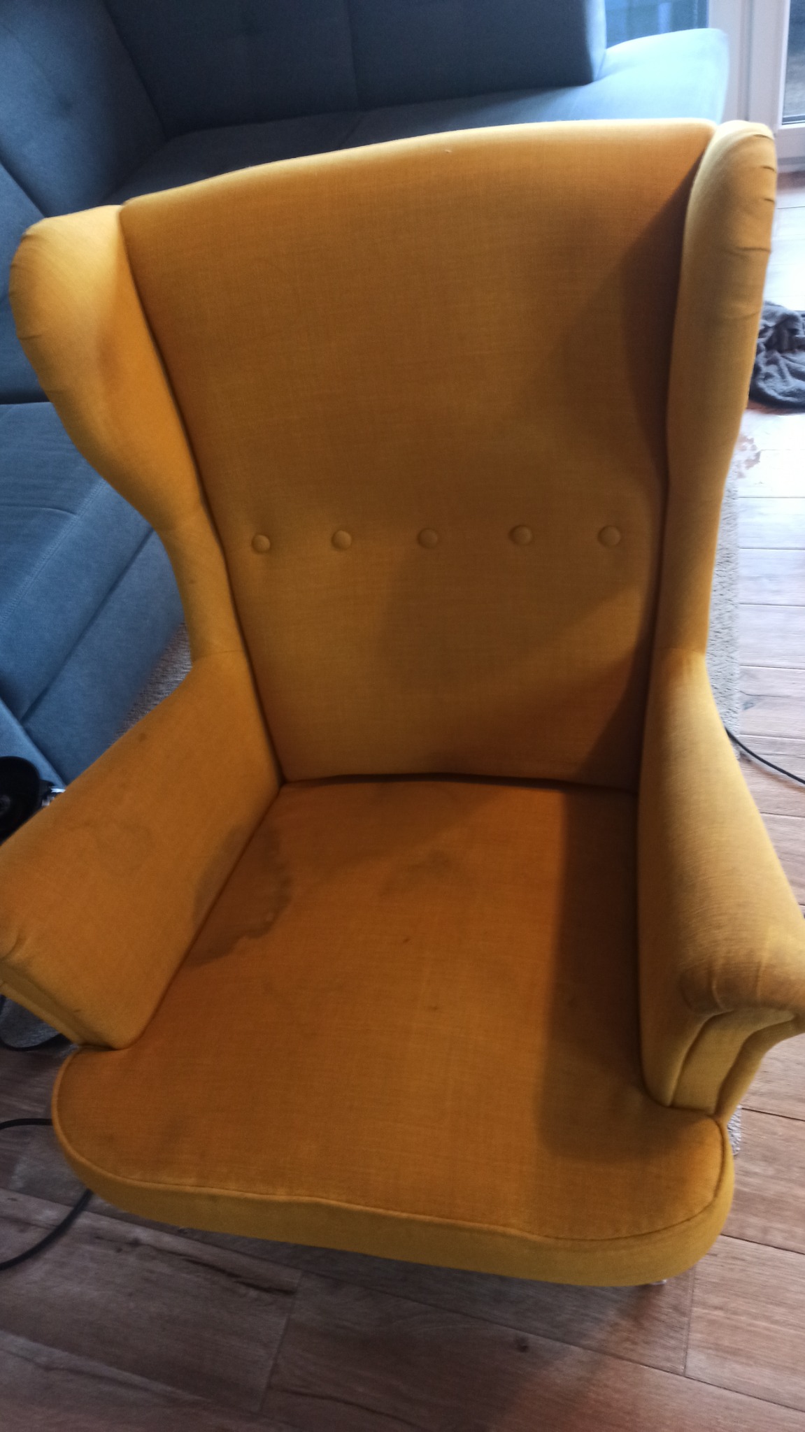 Żółty fotel typu uszak z widocznymi plamami na siedzisku, sfotografowany z góry, obok fragment niebieskiej sofy.