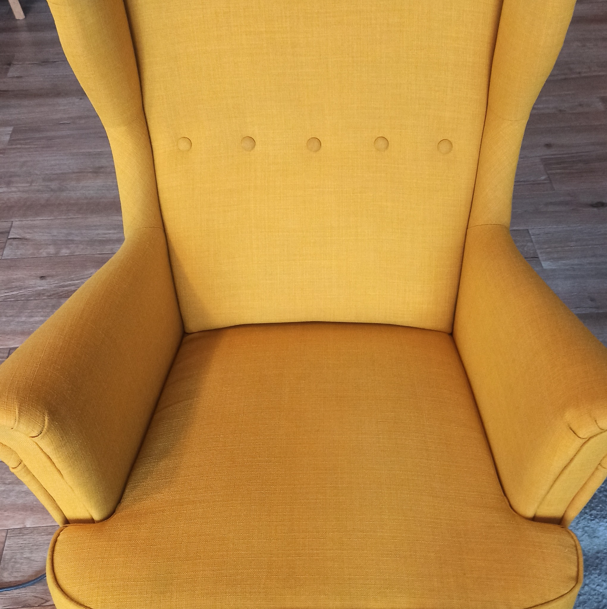Żółty fotel typu uszak z widoczną strukturą tkaniny i rzędem guzików na oparciu, ustawiony na panelach podłogowych.