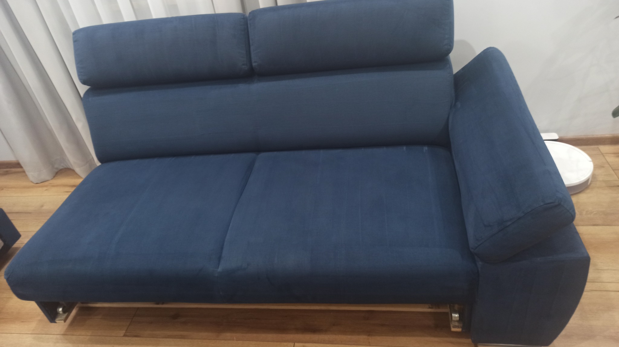 Ciemnoniebieska kanapa z regulowanymi zagłówkami i metalowymi nóżkami na drewnianej podłodze, obok widoczny biały robot sprzątający.