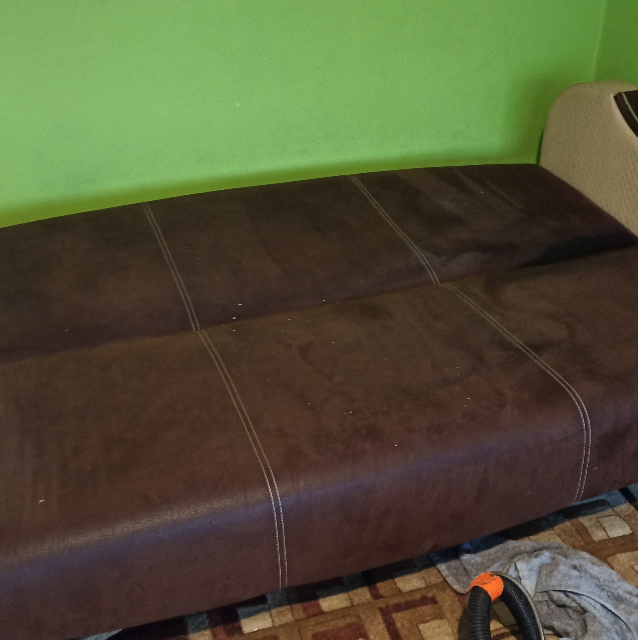 Brązowa kanapa z widocznymi plamami i śladami po czyszczeniu, obok leży szara ścierka i wąż od odkurzacza piorącego, zielona ściana w tle.