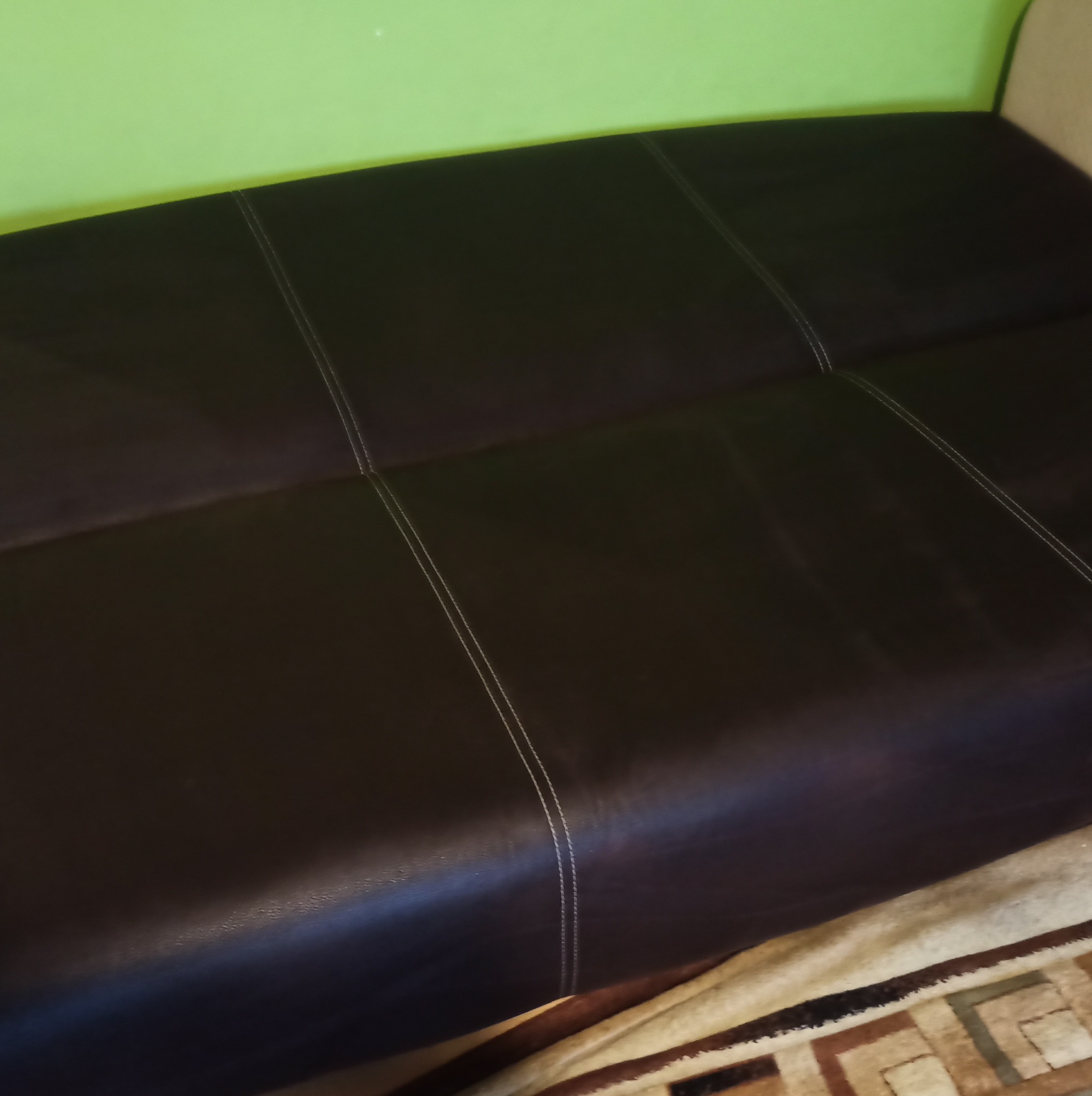 Fragment ciemnobrązowej kanapy ze szwami, widoczny fragment zielonej ściany i wzorzystego dywanu.