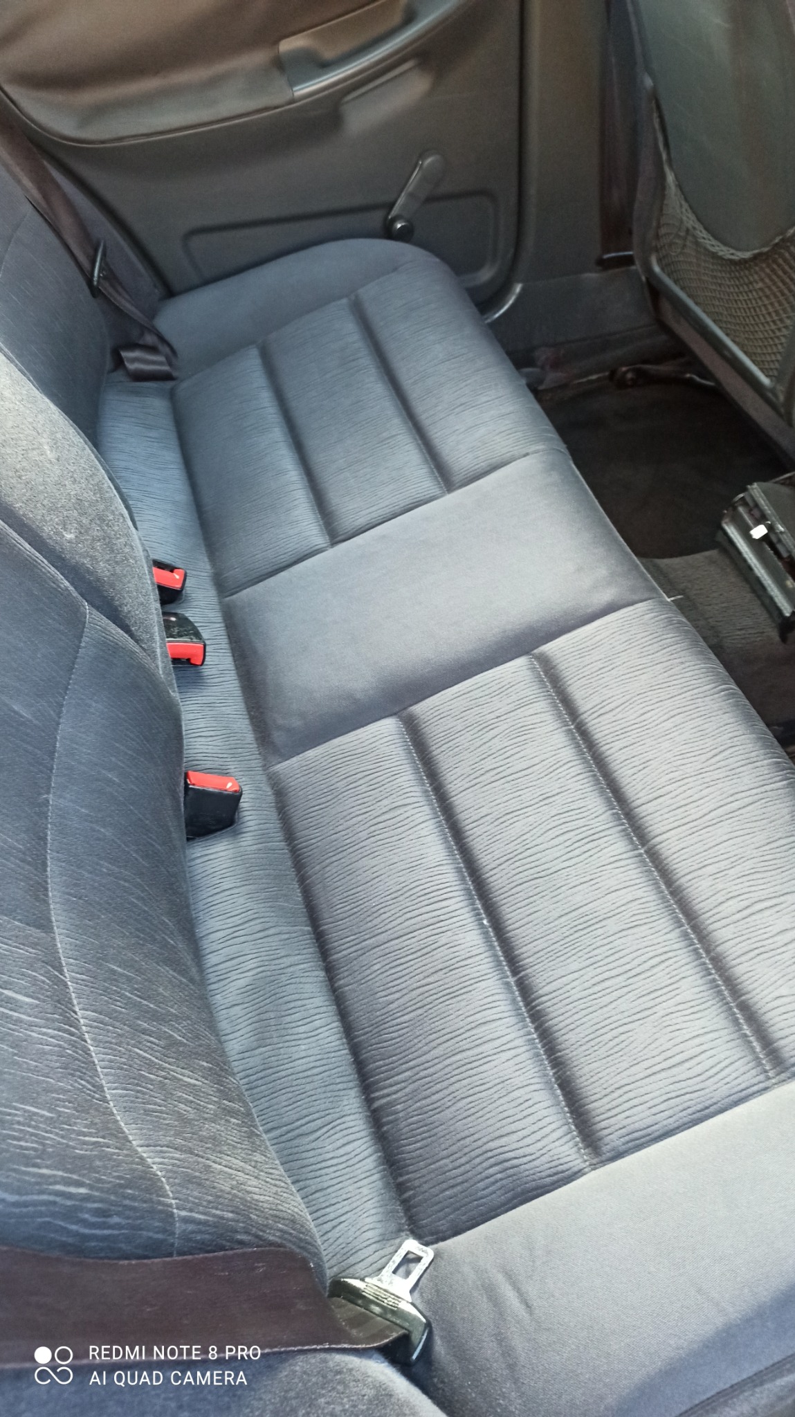 Szare, materiałowe siedzenie samochodowe z trzema zagłówkami i pasami bezpieczeństwa, widoczne ślady użytkowania.