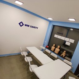 WM Safety sp. z o. o. - Sala szkoleniowa z białymi stołami i krzesłami, manekiny do nauki pierwszej pomocy widoczne w tle za oknem, logo WM Safety na ścianie.