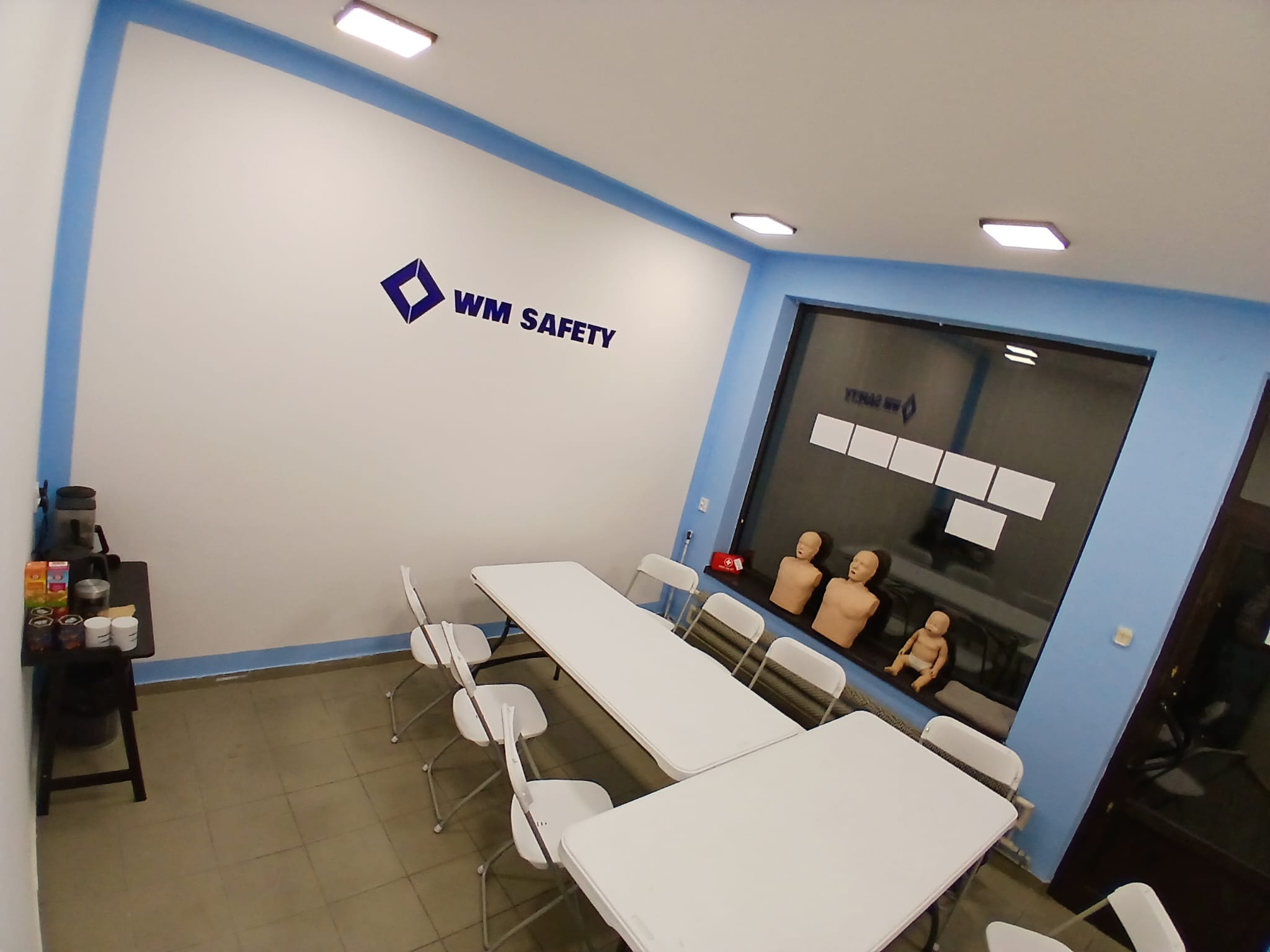 Sala szkoleniowa z białymi stołami i krzesłami, manekiny do nauki pierwszej pomocy widoczne w tle za oknem, logo WM Safety na ścianie.