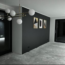 R-bud - Nowoczesne wnętrze w trakcie wykańczania: ciemnoszara ściana z abstrakcyjnymi obrazami w złotych ramach, surowa betonowa podłoga, designerska lampa sufitowa z białymi kulami, widoczne gniazdka...