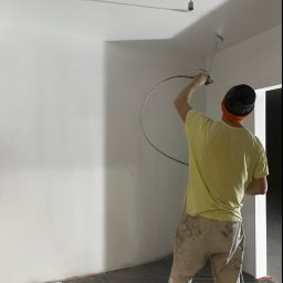 R-bud - Malowanie ścian natryskowo w pomieszczeniu z odkrytą żarówką, widoczne spodnie malarza z zabrudzeniami farby.