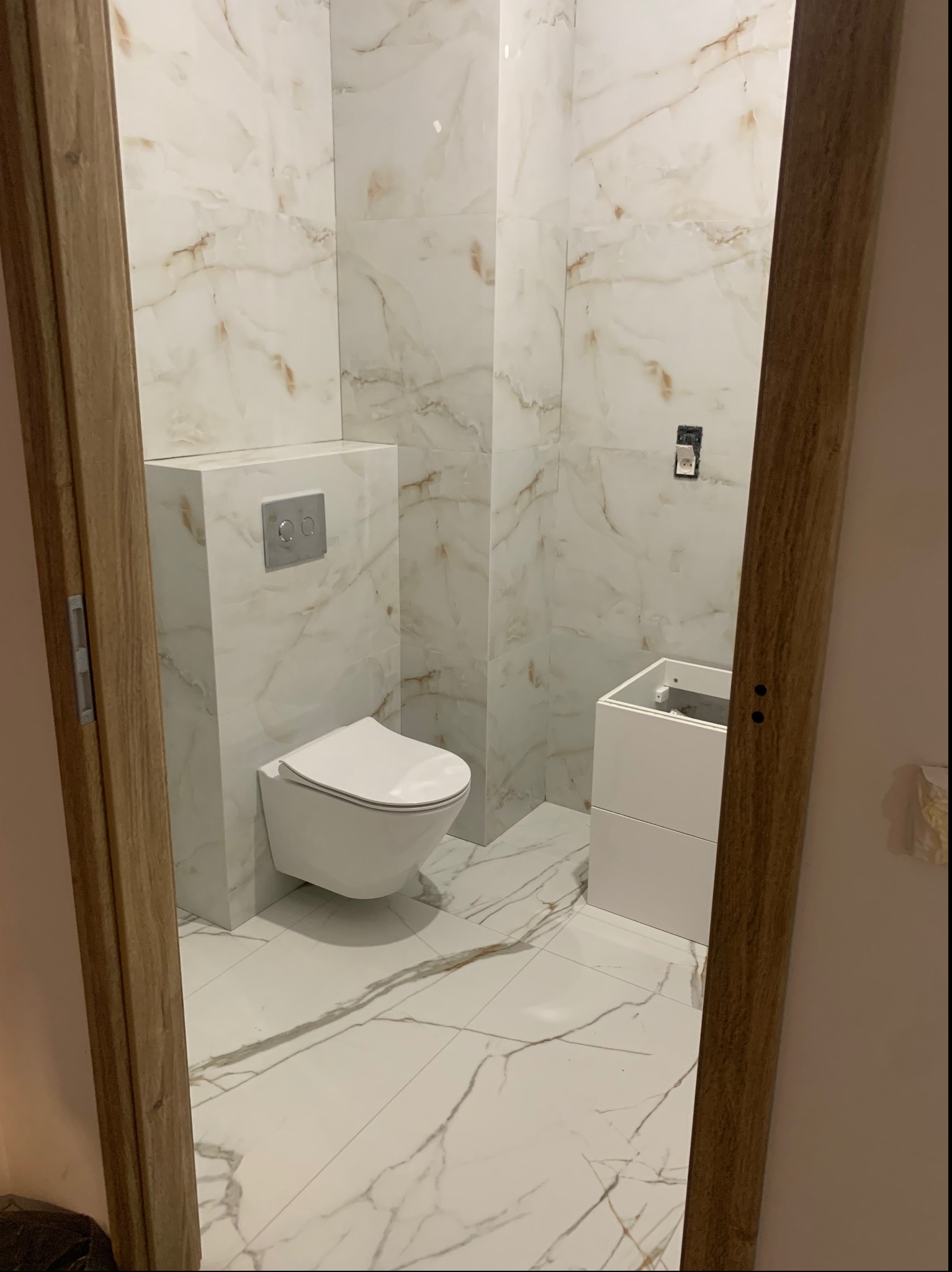 Nowoczesna łazienka z białymi płytkami imitującymi marmur, wisząca toaleta z chromowanym przyciskiem spłukującym i częściowo zamontowana biała szafka pod umywalkę, widok zza drewnianej ościeżnicy.