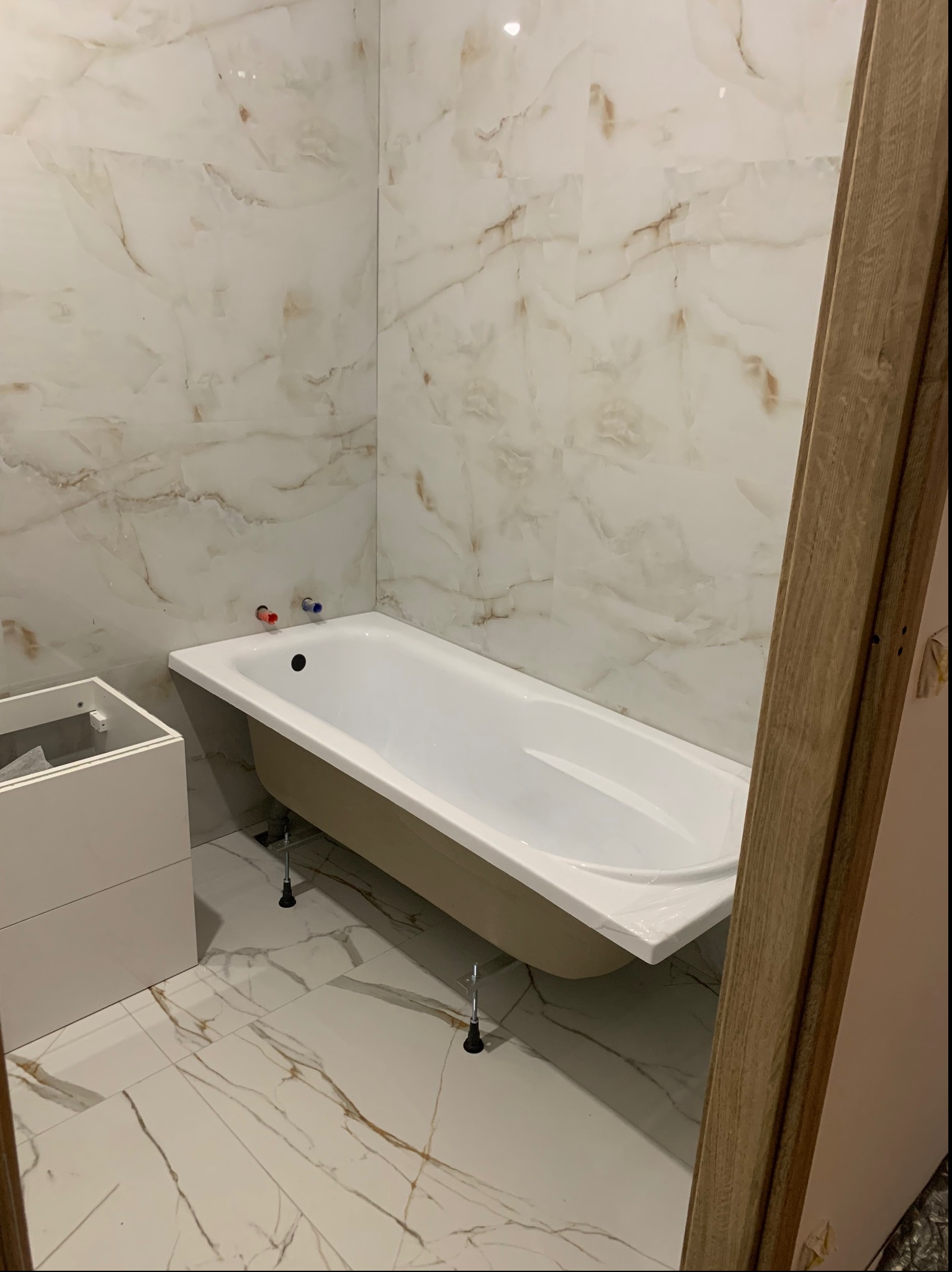 Biała wanna z zamontowanymi nóżkami, częściowo zabudowana, na tle marmurowych płytek ściennych i podłogowych, widoczny fragment ramy drzwi.