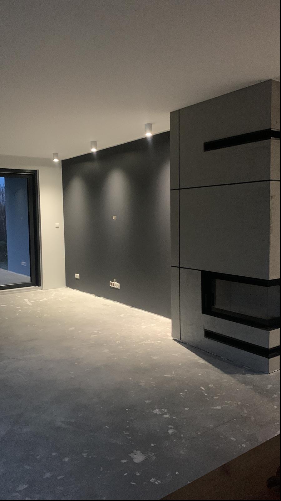 Surowe wnętrze w szarościach z minimalistycznym kominkiem w trakcie wykańczania, widoczne ślady po pracach na podłodze, czarna ściana z punktowym oświetleniem.