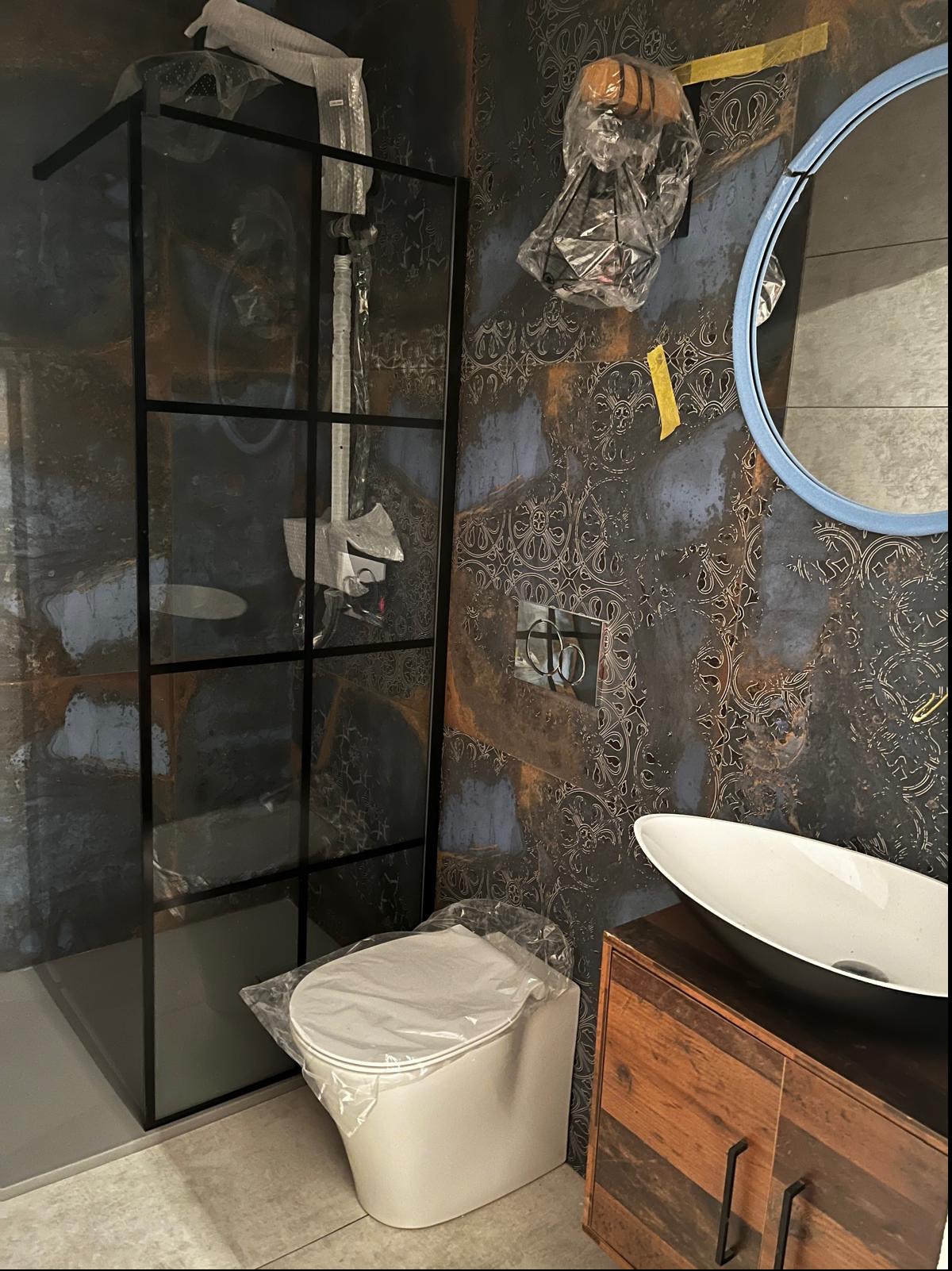 Nowa łazienka w trakcie wykańczania: kabina prysznicowa z czarnymi profilami, toaleta w folii ochronnej, umywalka nablatowa na drewnianej szafce, lustro w niebieskiej ramie i wzorzyste płytki...