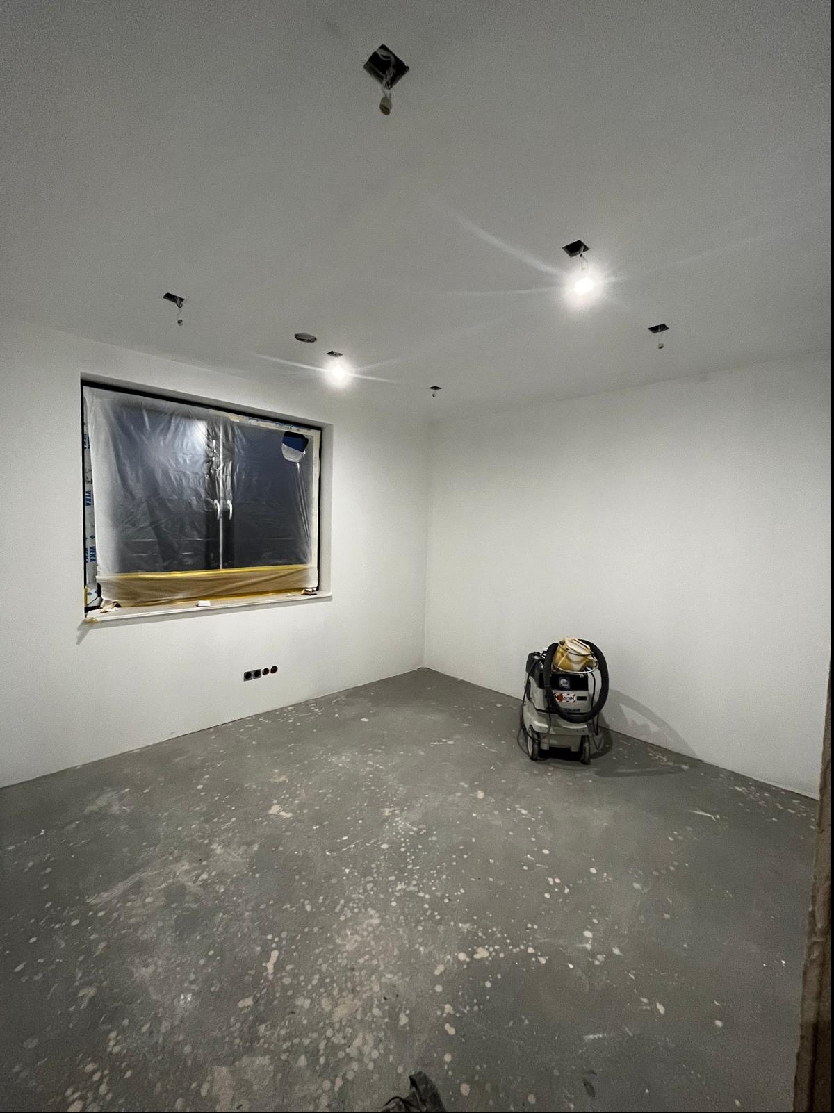 Pokój w trakcie prac wykończeniowych: świeżo pomalowane ściany na biało, zabezpieczone okno folią, widoczny odkurzacz przemysłowy i plamy na podłodze.