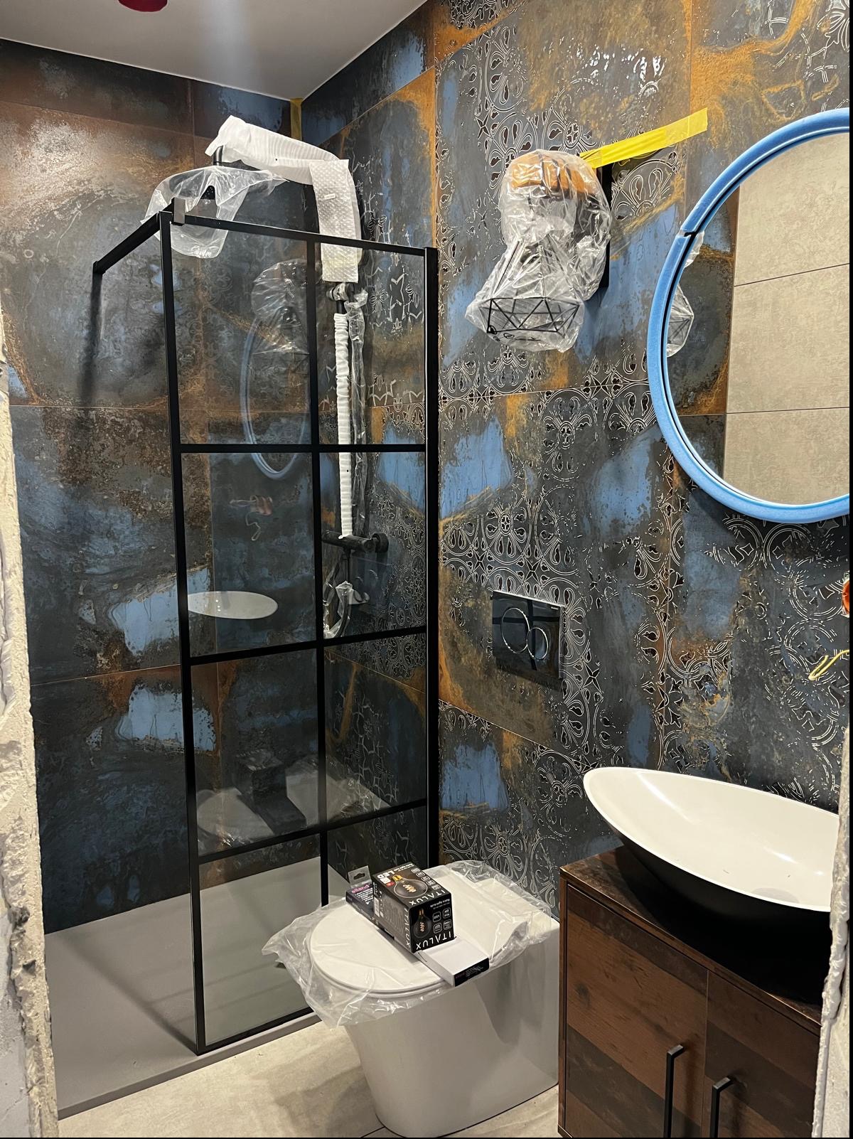 Łazienka w trakcie wykańczania: kabina prysznicowa z czarnymi profilami, umywalka nablatowa na drewnianej szafce, toaleta z pudełkiem żarówek na pokrywie, dekoracyjne płytki ścienne w odcieniach...